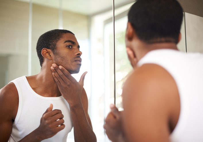 A young man with a neutral expression examines himself in the mirror.